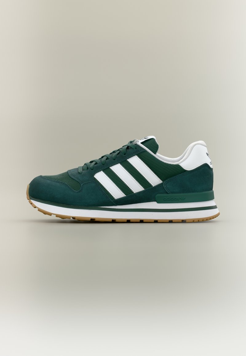 Mørkegrøn Adidas-sneaker med hvide striber, hvid hæftemærke bagpå, gummisål og snørebånd, vist i siddeprofil mod neutral baggrund.