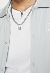Chemise boutonnée gris clair en tissu texturé ; chaînes en argent superposées avec un pendentif en forme de clé et un cadenas sur un t-shirt blanc en dessous.