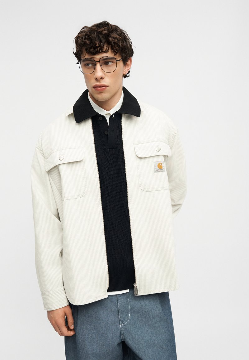 Junger Mann mit lockigem Haar und Brille, der eine cremefarbene Cordjacke über einem schwarzen Hemd mit Kragen und blauen Hosen trägt und nach links schaut.