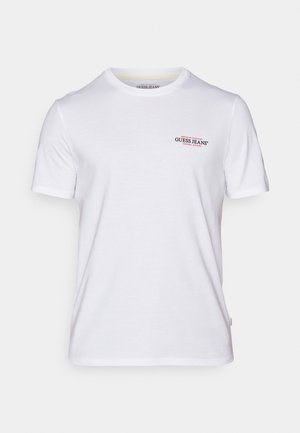 Baltas kokvilnas T-krekls ar apaļu kaklu, īsām piedurknēm un mazu sarkanu un zilu "GUESS JEANS" logo kreisajā krūšu pusē.
