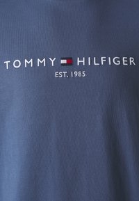 Navy blue cotton t-shirt featuring "TOMMY HILFIGER" embroidered in white, with a horizontal red and black stripe beneath the text. "EST. 1985" printed below.