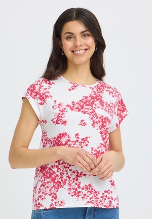 Donna sorridente con capelli scuri che indossa una camicetta bianca a maniche corte con motivi floreali rossi, in piedi con le mani intrecciate in modo rilassato.