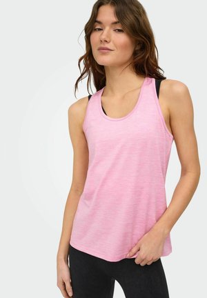 Ung kvinde iført en lyserød ærmeløs top over en sort sports-bh og sorte leggings, stående mod en ensartet lys baggrund.