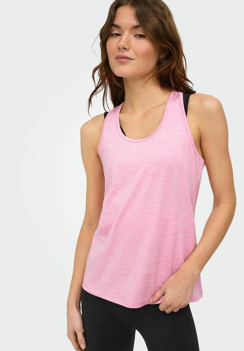 Młoda kobieta ubrana w różowy bezrękawnik na czarnym staniku sportowym, z czarnymi legginsami, stoi na tle jasnego, jednolitego tła.