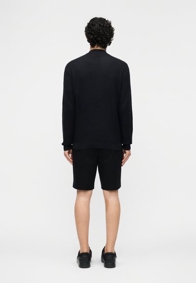 Άνδρας με σγουρά μαλλιά στέκεται με την πλάτη γυρισμένη, φορώντας μαύρο ριμπ πλεκτό πουλόβερ, μαύρο σορτς και μαύρα αθλητικά παπούτσια, σε μονόχρωμο λευκό φόντο.