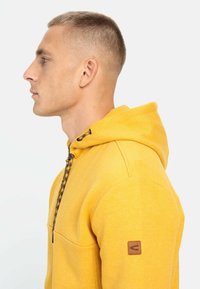 Gul hættebluse med frontlomme, tekstureret stof, sort snor og logopatch på venstre ærme. Sidesynsvinkel.