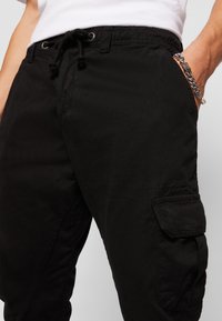 Černé cargo kalhoty z bavlny s elastickým pasem a šňůrkou na stažení. Mají boční kapsy a náprsní kapsu na stehně.