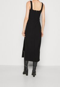 Robe noire sans manches avec un décolleté carré, une silhouette ajustée et une fente sur le côté. Fermeture éclair au dos. Texture de tissu lisse.