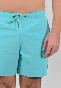 Pantaloni da bagno turchesi con vita elastica, chiusura con cordoncino e logo ricamato sulla gamba destra. Tessuto liscio e leggero.