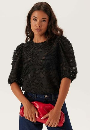 Svart texturerad blus med puffärmar och blommönster, i kombination med en röd clutch och denimjeans. Slät tyg med lagerdesign detaljer.