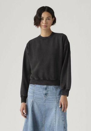 Kvinde med kort mørkt hår iført en sort sweatshirt med rund hals og en lyseblå cowboynederdel, der står mod en ensfarvet baggrund.