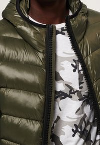 Chaqueta acolchada brillante de color verde oliva con cremallera frontal y capucha, sobrepuesta a una camiseta gris claro con un patrón de camuflaje en negro y gris.