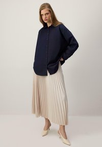 Femme portant une chemise boutonnée oversize bleu marine, une jupe midi plissée crème et des talons pointus crème, debout contre un fond uni.