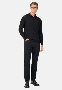 Camisa polo negra de manga larga con cuello, combinada con pantalones negros y zapatos de vestir negros. El modelo lleva gafas y está de pie con las manos en los bolsillos.