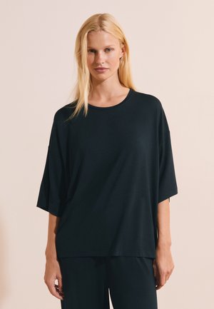 Blonde woman wearing loose black short-sleeve top and matching black pants, standing against a plain beige background.