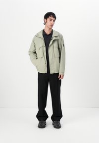 Light green hooded jacket with four front pockets, layered over a black polo shirt, paired with black wide-leg pants and dark sneakers.
