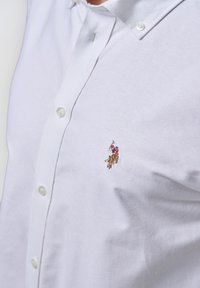 Camicia bianca con bottoni, caratterizzata da una trama sottile e un piccolo logo ricamato di un giocatore di polo sul petto. Colletto standard con bottoni di plastica.