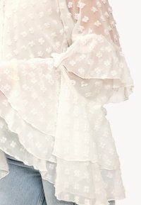 Blouse blanche en tissu léger et texturé, présentant un motif floral à pois, des manches à volants et un détail de ourlet superposé.