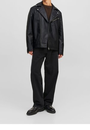 Henkilö pukeutunut mustaan nahkaiseen moottoripajakkiin, tummanruskeaan villapaitaan, löysiin mustiin housuihin ja mustiin nahkakenkiin, yksinkertaisen harmaan taustan edessä.