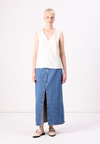Haut sans manches beige clair avec un col en V, associé à une longue jupe en denim bleue avec une fente devant. Les chaussures sont de couleur claire avec des lanières.
