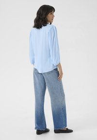 Blouse bleue, texturée, à manches longues et manches bouffantes, associée à un jean large bleu clair. Des chaussures plates noires complètent la tenue.