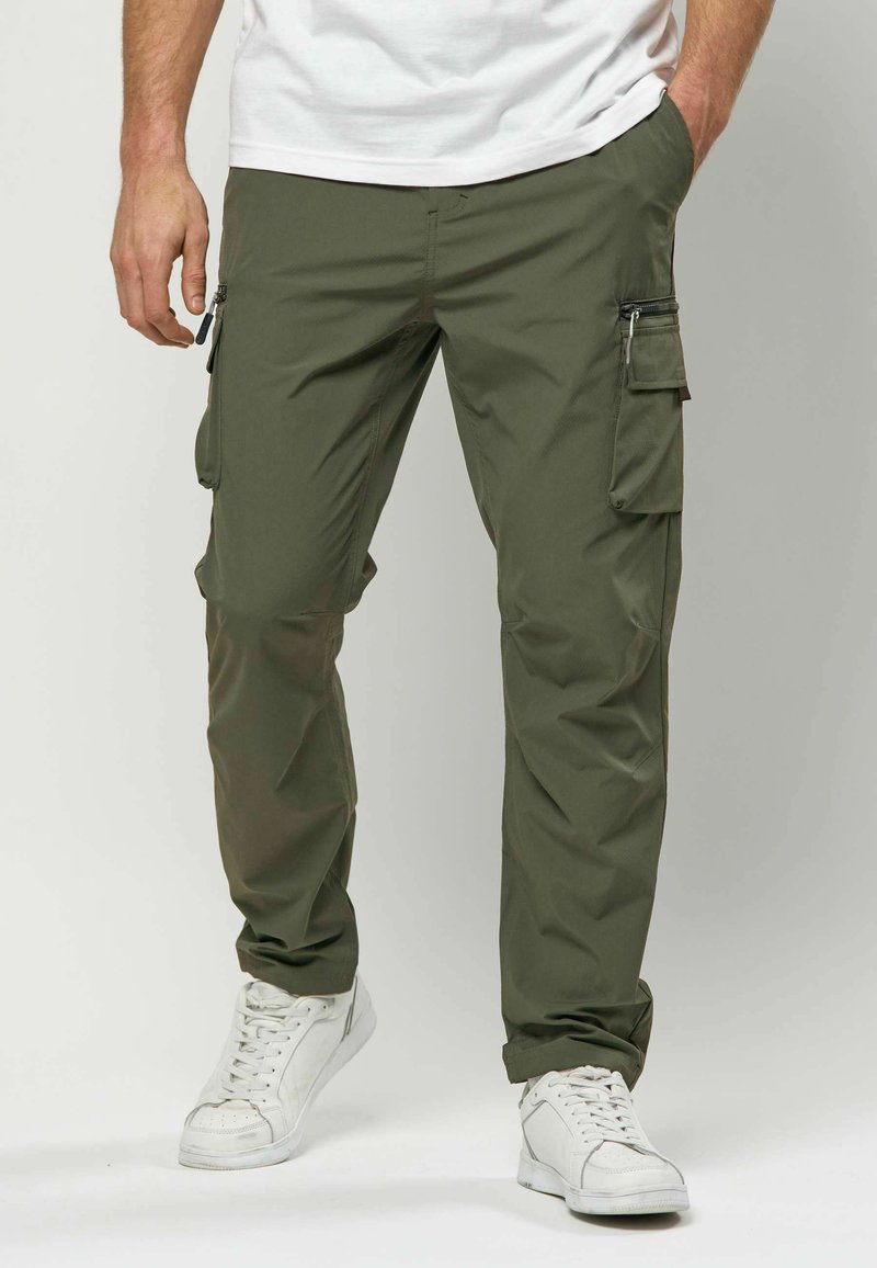 Homme portant un pantalon cargo vert olive avec des poches latérales et des baskets blanches, debout avec une main dans la poche et l'autre détendue.