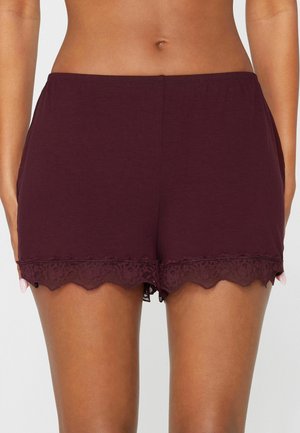 Femme portant un short violet foncé bordé de dentelle, montrant le ventre et le haut des cuisses sur un fond uni.