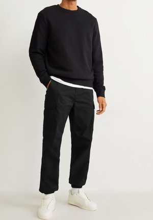 Homme portant un sweat-shirt noir, un pantalon cargo noir, des baskets blanches et des chaussettes blanches, debout avec une main dans la poche devant un fond blanc.