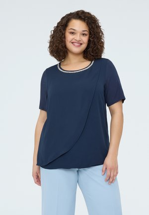 Smilende kvinde med krøllet hår iført en mørkeblå kortærmet top med dekorativ halsudskæring og lyseblå bukser, stående foran en ensfarvet baggrund.