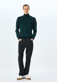 Mörkgrön turtleneck-tröja med ribbad textur vid axlarna och bröstet, kombinerad med mörka denimjeans och vita sneakers.
