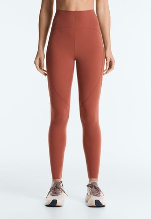 Woman wearing rust-colored high-waist leggings and white sneakers, standing against a plain light background.