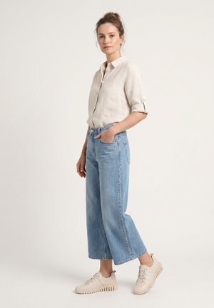 Vrouw staand, draag een beige overhemd met knopen en opgerolde mouwen, lichtblauwe brede jeans en beige veterschoenen voor dagelijks gebruik.