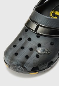 Black clogs with a textured surface, ventilation holes, and a decorative silver logo. Features a glossy black strap and rounded design.