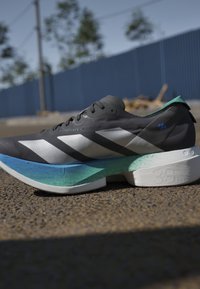 Scarpa da corsa bianca e nera con suola a gradiente blu e verde, posizionata sull’asfalto con una recinzione blu sfocata e alberi sullo sfondo.