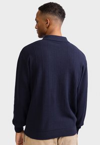 Άνδρας με πλάτη που φοράει μπλε ναυτικό υφασμάτινο πουλόβερ με μακριά μανίκια και κάθετες ρίγες, καθώς και μπεζ παντελόνι, μπροστά σε απλό φόντο.