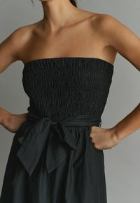 Mustaa olkaimetonta mekkoa, jossa on kirjailtu vartalo, vyötäröä korostava leikkaus ja rusettiyksityiskohta. Valmistettu kevyestä kankaasta, jossa on liehuva, rento helma.