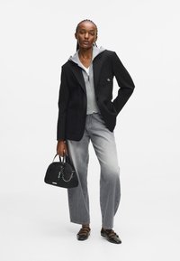 Čierny blazer cez šedú mikinu na zips, v kombinácii so šedým tielkom, svetlými džínsami a čiernymi topánkami. Drží malú čiernu kabelku.