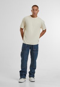 Cream-colored short-sleeve t-shirt with a relaxed fit, paired with blue cargo jeans with side pockets. The shoes are white athletic sneakers.