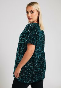 Top di paillettes nera e teal con un fit morbido, maniche corte e orlo arrotondato. La texture è lucida e riflettente.
