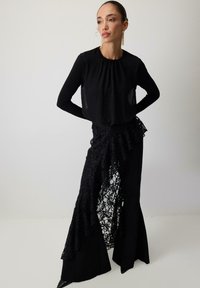 Melna gara piedurknēm blūze ar plisētu kakla izgriezumu, kombinēta ar piegulīgu melnu svārku ar slāņainām mežģīņu detaļām un izplūdušu siluetu.