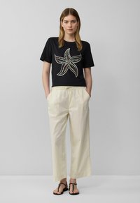 Zwarte cropped t-shirt met een bloemenpatroon, gecombineerd met losse beige broeken en zwarte platte sandalen. Minimalistisch ontwerp en een ontspannen pasvorm.