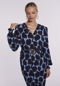 Niebieska i czarna sukienka w wzory z dekoltem w serek, długimi bufkami i talią podkreśloną. Posiada ozdobny pasek z okrągłą klamrą.