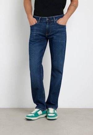 Jeans in denim scuro con taglio dritto, caratterizzati da tasche frontali e chiusura con bottone. Indossati con sneaker verdi e bianche.