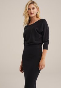 Černý pletený top s širokým výstřihem a dlouhými rukávy, kombinovaný s přiléhavou černou sukní. Hladká textura, minimalistický design, bez viditelných vzorů.