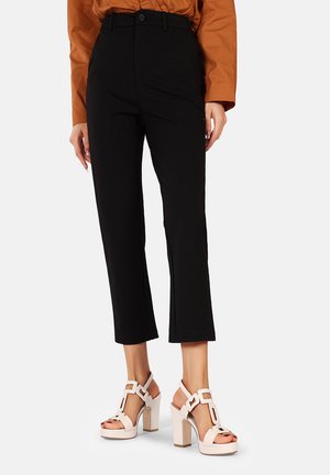 Pantaloni neri su misura con una linea dritta, abbinati a sandali beige con tacco a blocco che presentano un dettaglio a strappo geometrico.