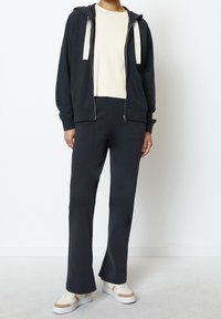 Felpa nera con zip e cordini grigio chiaro, abbinata a una maglietta a maniche corte cremisi a coste e pantaloni neri a campana. Sneakers bianche e beige.