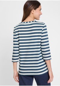 Eine Frau lächelt und trägt ein navyblaues und weißes, horizontal gestreiftes Shirt mit dreiviertel Ärmeln, zu sehen von hinten.