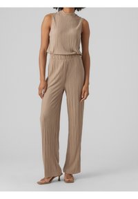 Beżowy kombinezon bez rękawów z plisowaniem, wysokim dekoltem i szerokimi nogawkami, noszony z beżowymi sandałami na obcasie.