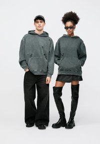 Dvaja ľudia nosia oversized sivé mikiny z mäkkej látky, s vreckami a opotrebovanými detailmi. Muž má na sebe čierne nohavice a žena sukňu a vysoké čierne čižmy.