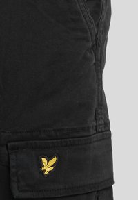 Black cargo pants with a textured finish, featuring side pockets and a small yellow embroidered eagle logo on the side pocket flap.
